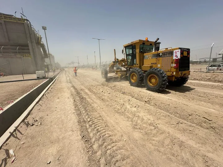 A motor grader working on an unpaved route beside industrial structures.