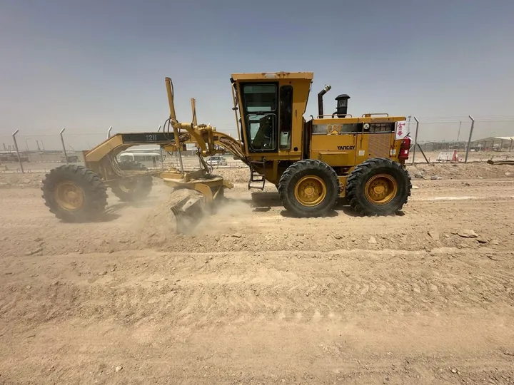 A yellow motor grader operating on an unpaved site surface.