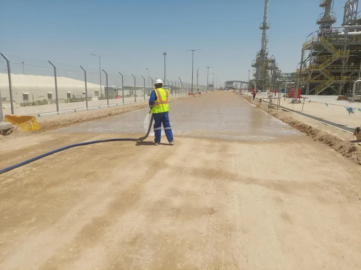 A worker using a hose to wet an unpaved road surface.