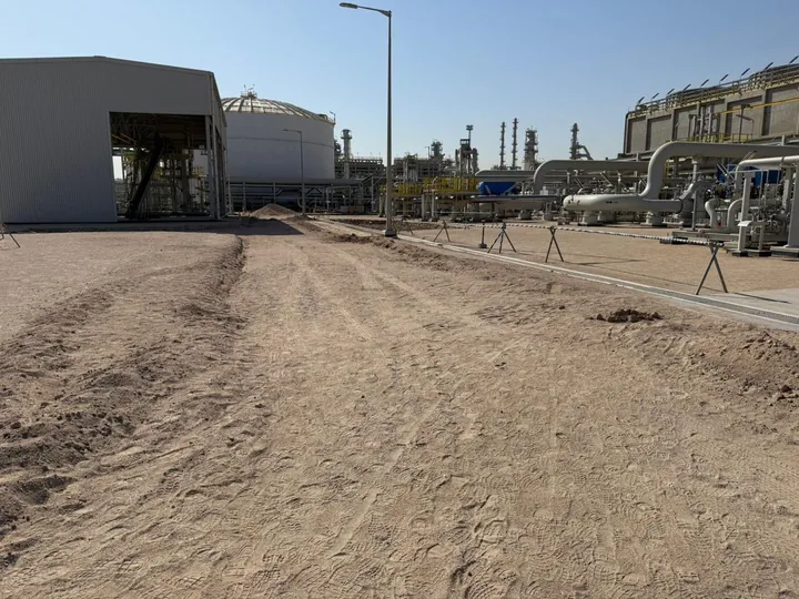 A graded unpaved route beside industrial pipework and equipment.