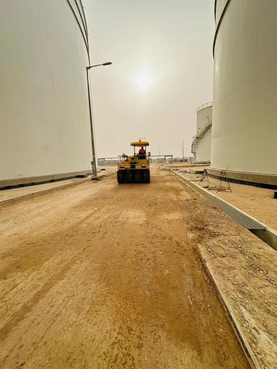 A road roller on an unpaved route between large storage tanks.